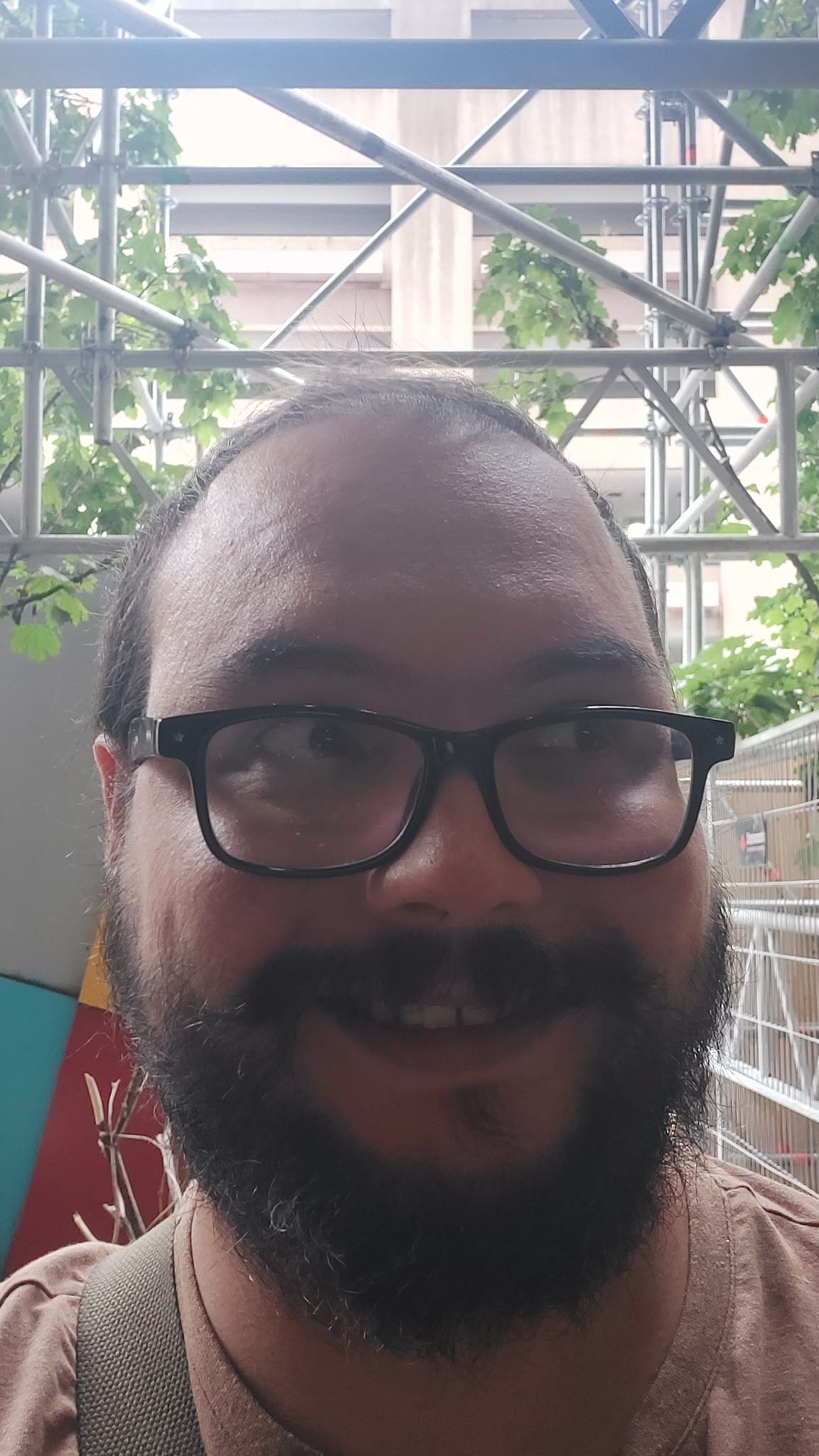 A man short hair and bushy facial hair stands in front of construction scaffolding looking towards the side